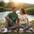 Couple in forest green and natural Time by the River t-shirts with matching track pants showing design on lower legs by river at sunset